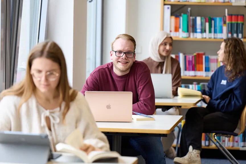 Ein Studierender sitzt an einem Arbeitsplatz in einer Bibliothek und lächelt in die Kamera