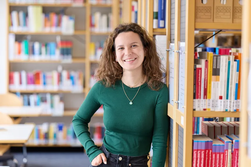 Eine Studierende steht an einem Bücherregal und lächelt