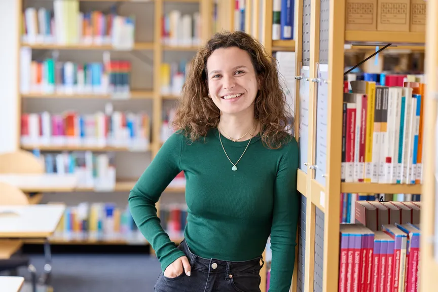 Eine Studierende steht an einem Bücherregal und lächelt