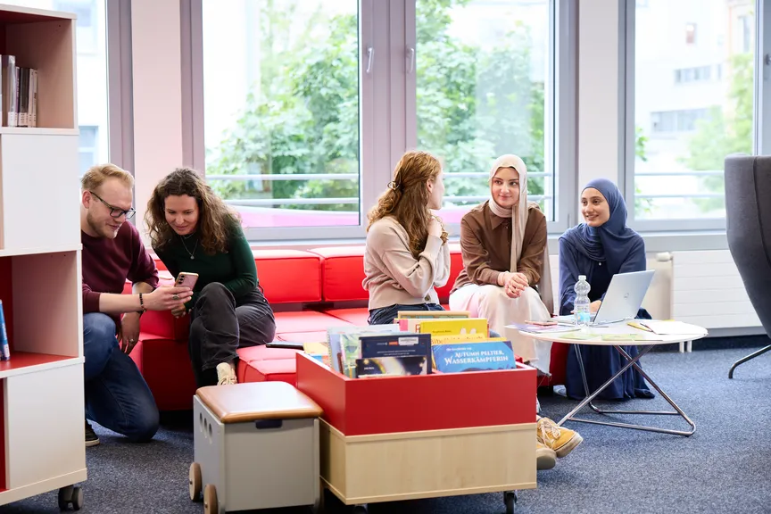 Einige Studierende sitzen auf einer Sitzmöglichkeit in einer Bibliothek und reden miteinander