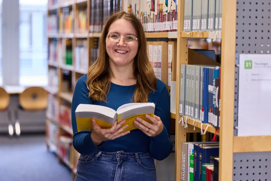 Eine Studierende steht an einem Bücherregal, hält ein Buch in der Hand und lächelt