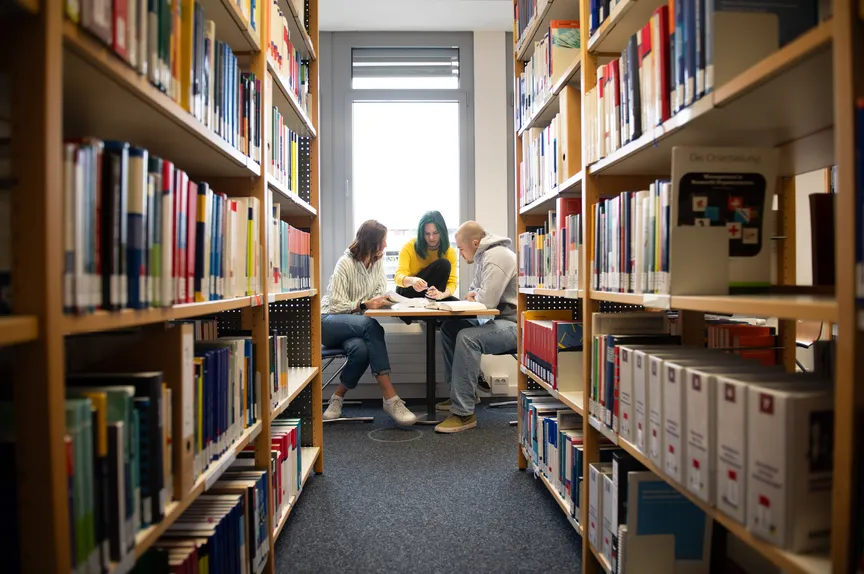 Einige Studierende sitzen in der Bibliothek an einem Arbeitsplatz vor Bücherregalen