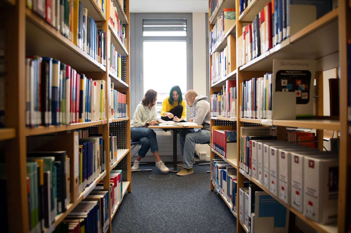 Einige Studierende sitzen in der Bibliothek an einem Arbeitsplatz vor Bücherregalen