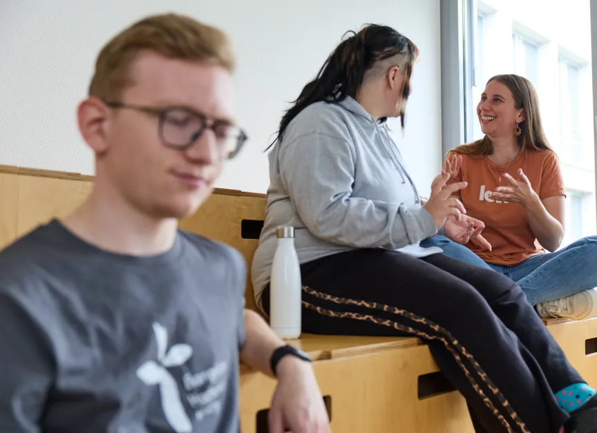 Einige Studierende sitzen auf einer hölzernen Sitzgelegeneheit und unterhalten sich