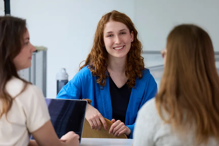 Eine Studierende sitzt in einer Gruppe Studierender in einem Seminar und lächelt in die Kamera