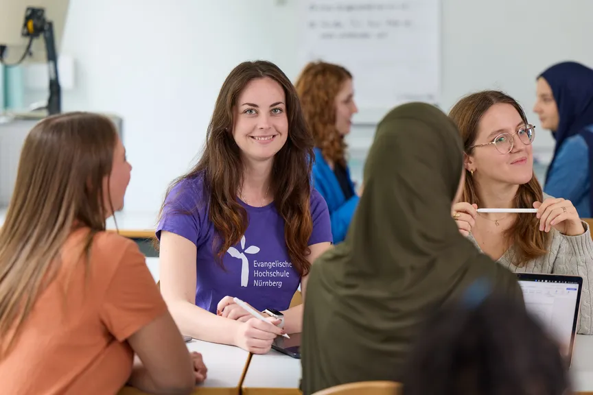 Eine Studierende sitzt in einer Gruppe Studierender in einem Seminar und lächelt in die Kamera