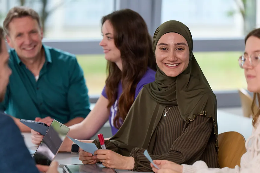 Eine Studierende sitzt in einer Gruppe Studierender in einem Seminar und lächelt in die Kamera