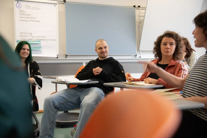 Einige Studierende sitzen auf modernen Stühlen in einem Seminarraum und unterhält sich