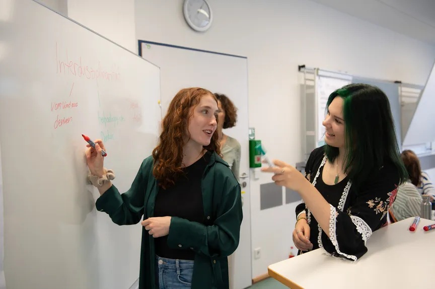 Zwei Studierende schreiben mit Markern auf eine Tafel und diskutieren