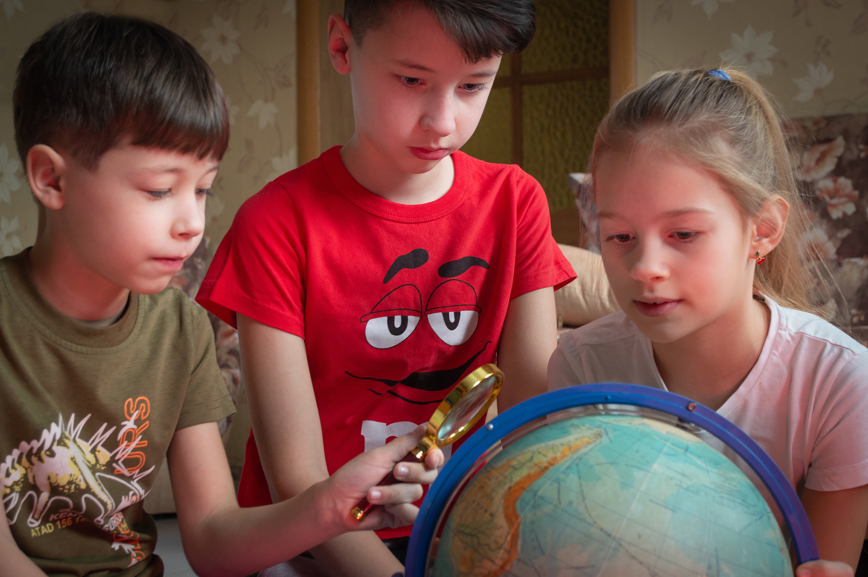 Drei Kinder im Grundschulalter schauen auf einen Globus. Ein Kind hält eine Lupe in der Hand
