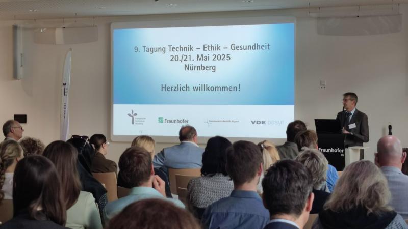 Tagung technik-Ethik-Gesundheit im Konferenzsaal mit Präsentation, am Rednerpult Bernhard Opolony, Amtschef im Staatsministerium für Gesundheit, Pflege und Prävention