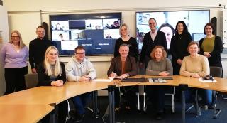 Elf erwachsene Männer und Frauen sitzen an einem Konferenztisch, im Hintergrund zwei große Bildschirme mit Zoom-Teilnehmenden