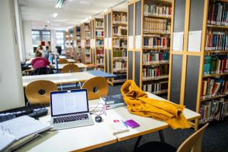 Bibliothek, Bücherregale, Tische, Laptop und Jacke auf Tisch, im Hintergrund Studierende an Tischen